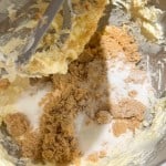 A stand mixer bowl containing creamed butter, white sugar, and brown sugar, with a paddle attachment partially coated in the mixture.