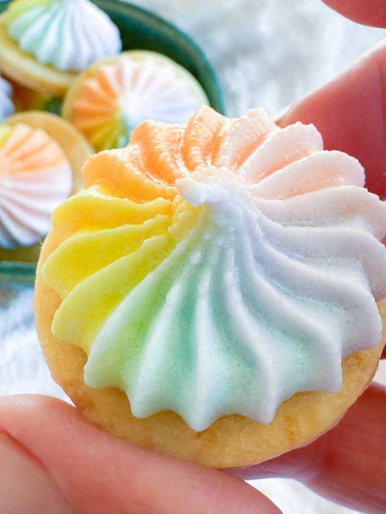 A hand holding a round cookie topped with a swirl of rainbow-colored meringue, with more similar cookies in a bowl in the background.
