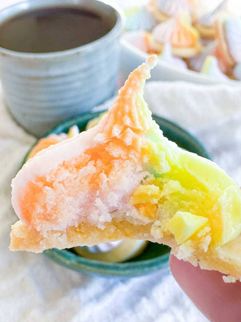 A hand holding a bite-sized, rainbow-colored meringue cookie, with a cup of coffee in the background.