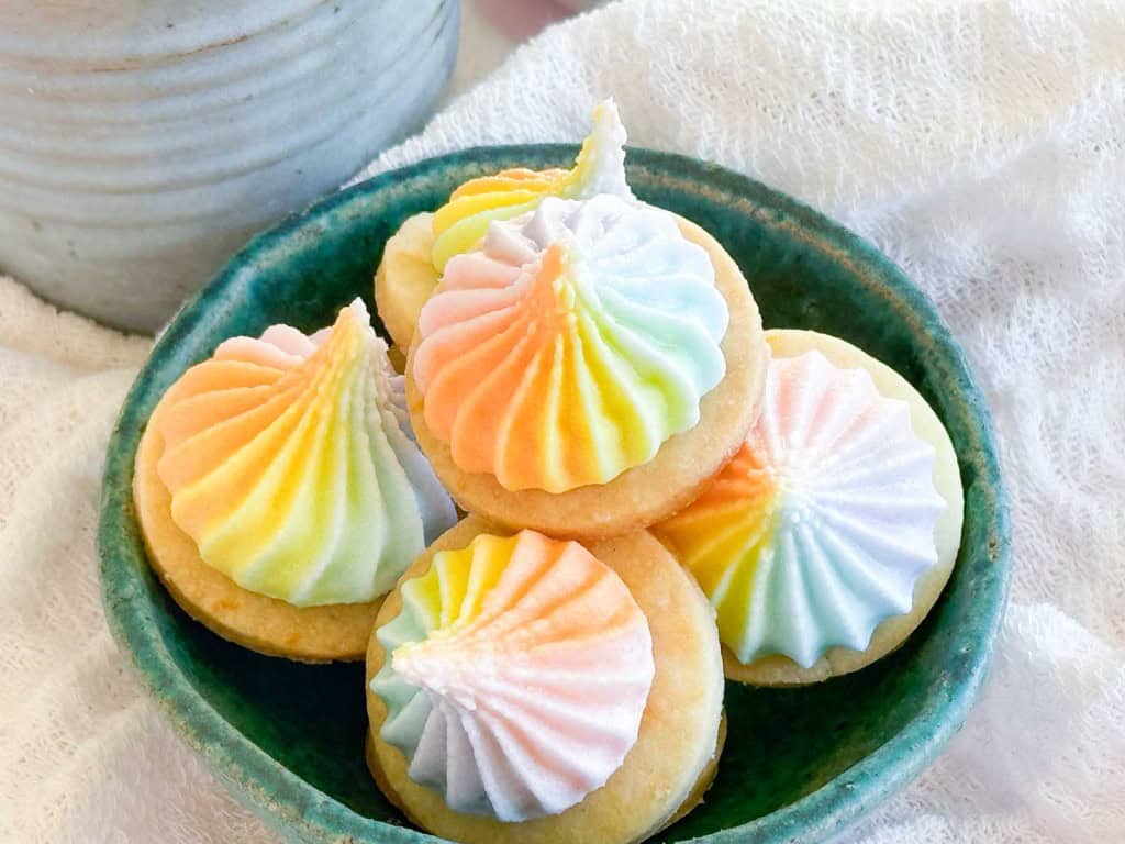 A small green bowl filled with round shortbread cookies topped with swirled, rainbow-colored meringue kisses.