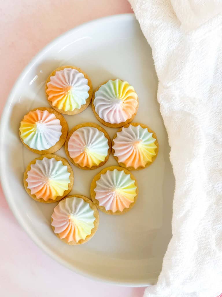 Seven Iced Gems cookies on a white plate, each topped with a swirl of pastel rainbow frosting.