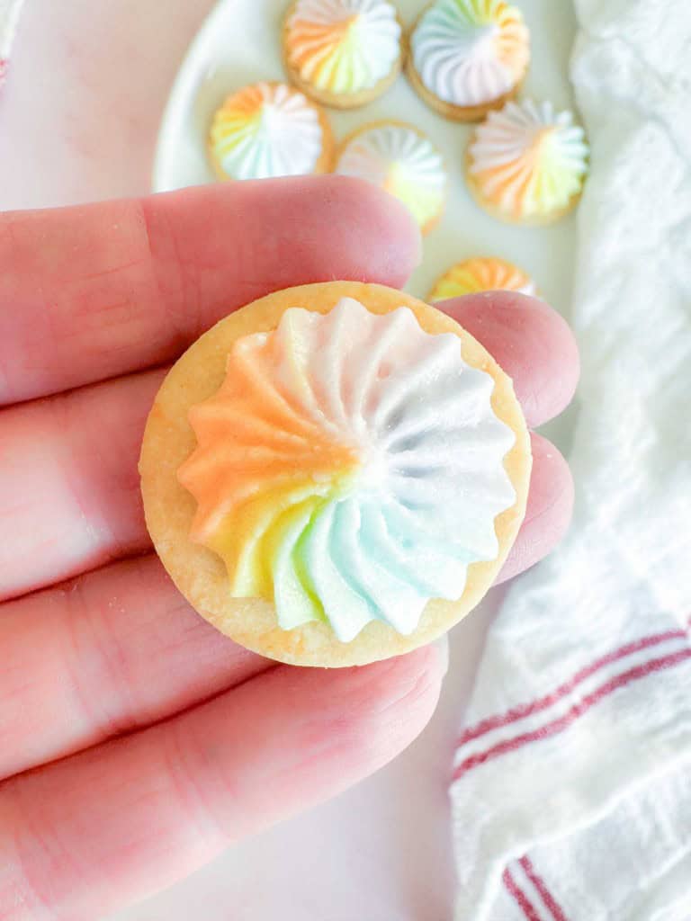 A hand holding a round Iced Gem topped with a swirl of rainbow-colored icing, with more cookies on a plate in the background.