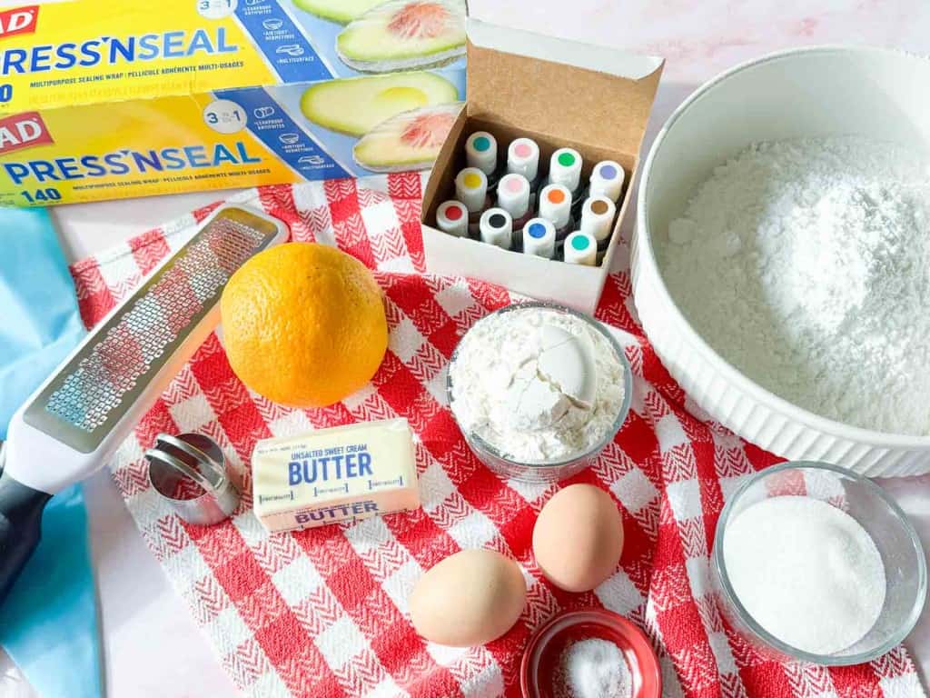 Baking ingredients for Iced Gems, including Press&rsquo;n Seal, food coloring, an orange, butter, eggs, flour, sugar, salt, and a grater on a red-and-white checkered cloth.