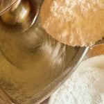 A spoonful of white flour being poured into a metal stand mixer bowl to prepare the base for Iced Gems.