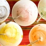 Five bowls of pastel-colored frosting, reminiscent of iced gems, each with a metal spoon, arranged on a striped cloth.
