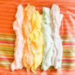 Five vertical rows of pastel-colored Iced Gems frosting&mdash;pink, orange, yellow, mint, and white&mdash;laid side by side on a sheet of plastic wrap.