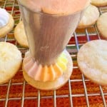 A piping bag frosts a round Iced Gem cookie with a multi-colored swirl of icing on a wire cooling rack.