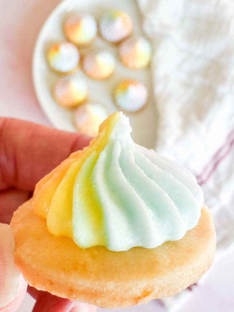 A hand holding a round Iced Gem topped with a swirl of rainbow-colored icing, with a plate of similar cookies in the background.