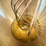 A metal whisk inside a mixing bowl containing raw egg whites for Iced Gems.