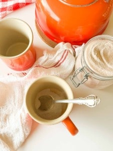 Two orange mugs, one with a silver spoon, beside a jar of beige powder and an orange kettle.