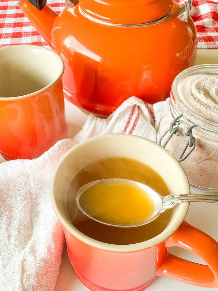 A spoonful of broth over a mug of soup, with an orange kettle and a jar of powder in the background.