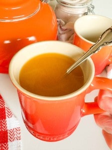 A hand holding an orange mug filled with broth, with a silver spoon inside, beside an orange kettle and another mug.