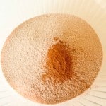 A glass bowl containing a mound of light brown flour with a small pile of cinnamon in the center.