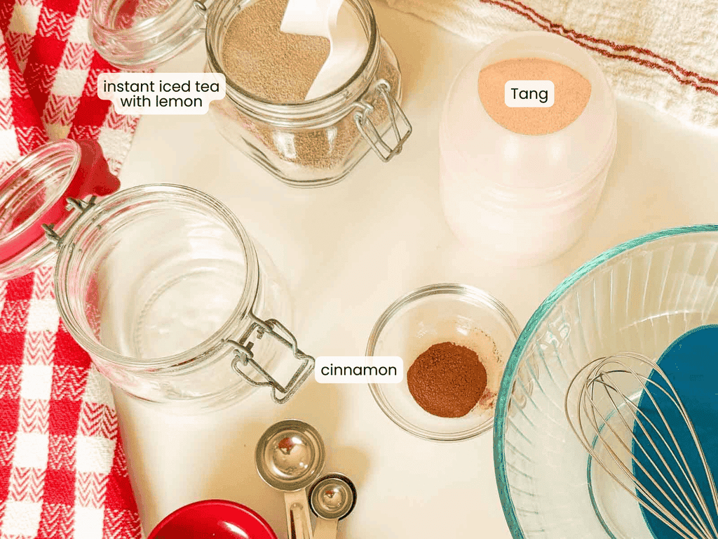 Ingredients for a drink mix including instant iced tea with lemon, Tang, and cinnamon, shown with measuring spoons, jars, and a whisk.