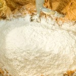Flour being added to a mixing bowl containing cookie dough with an electric mixer attachment.