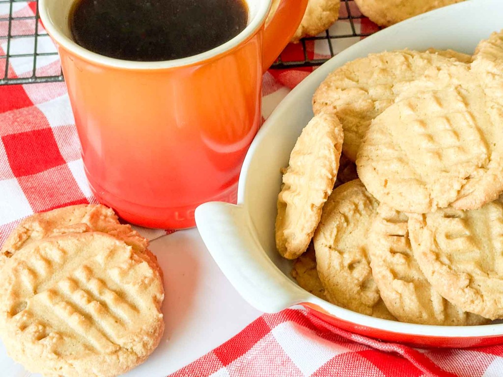 A mug of black coffee next to a white bowl filled with peanut butter cookies on a red and white checkered tablecloth.