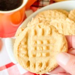 A hand holding a peanut butter cookie with a cross-hatch pattern, with a plate of cookies and an orange mug in the background.