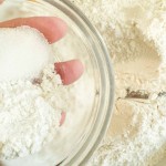 A glass bowl containing white sugar and flour held over a larger pile of flour.