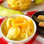 A small bowl of sliced pickled banana peppers, with a glass jar of pickled peppers and whole peppers on a red and white checkered cloth.